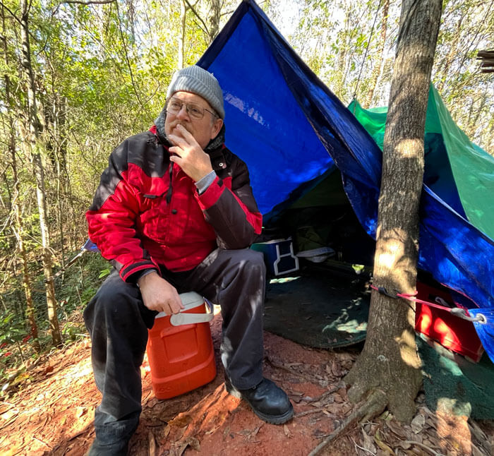 man sitting on a cooler in front of a tent in the woods